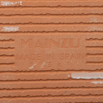 O ladrilho de terracota exibe em relevo o texto `MAINZU MADE IN SPAIN` em meio a ranhuras horizontais, sugerindo detalhes de ori