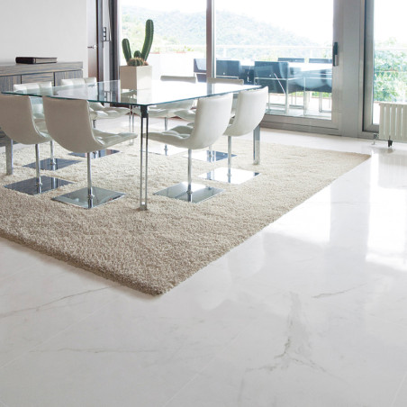 A dining area features a glass table with white chairs on a beige rug, surrounded by light marble flooring and large windows wit