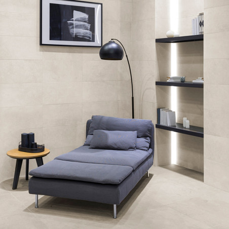 Een grijze chaise longue staat in een minimalistische kamer met beige betegelde muren. Hij staat onder een zwarte vloerlamp, naa