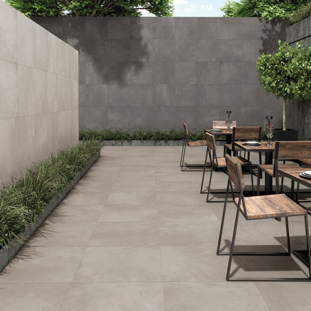 Buitenterras met grijze tegels. houten stoelen en tafels staan ​​nonchalant opgesteld. Weelderig groen omlijnt de muren en creëe