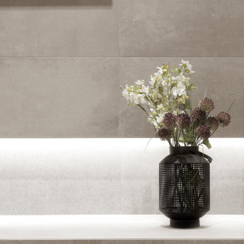 A black vase holds white and purple flowers, sitting on a light stone ledge against a textured beige tiled wall.