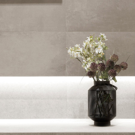 Um vaso preto contém flores brancas e roxas, apoiado em uma borda de pedra clara contra uma parede de azulejos bege texturizados