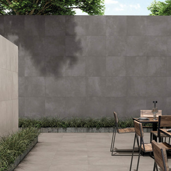 A minimalist concrete patio features wooden chairs around a metal table, bordered by a gray tiled wall and green grass under tre