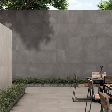 Een minimalistische betonnen patio met houten stoelen rond een metalen tafel, begrensd door een grijs betegelde muur en groen gr