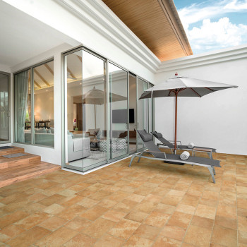 Twee grijze loungestoelen rusten onder een grote parasol op een lichtbruin betegeld terras. Aangrenzend kijkt een moderne kamer 