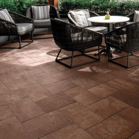 Brown square tiles form a patio surface. Wicker chairs surround a small white table, set in an outdoor area with lush greenery. Brown square tiles form a patio surface. Wicker chairs surround a small white table, set in an outdoor area with lush greenery.