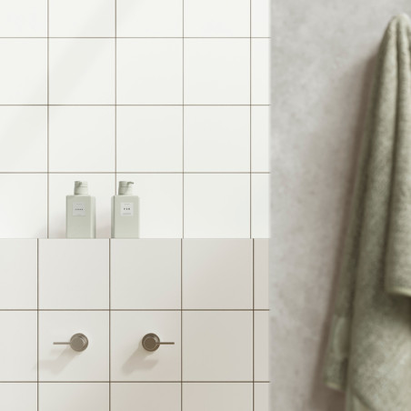 White tiles form a bathroom wall with two cylindrical handles below two bottles labeled `HAIR` and `BODY.` A green towel hangs n White tiles form a bathroom wall with two cylindrical handles below two bottles labeled `HAIR` and `BODY.` A green towel hangs n