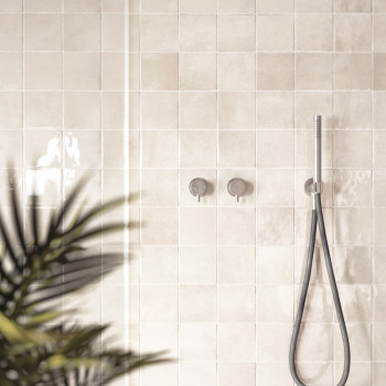 Azulejos bege brilhantes revestem a parede do banheiro, refletindo a luz. Um chuveiro e controles estão montados, com uma planta