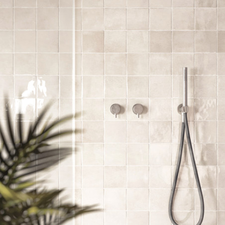 Azulejos bege brilhantes revestem a parede do banheiro, refletindo a luz. Um chuveiro e controles estão montados, com uma planta