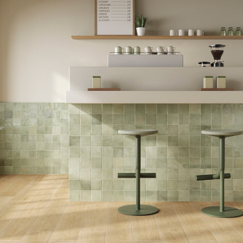 Deux tabourets verts se trouvent devant un comptoir de bar avec un mur de carreaux texturés dans les tons blancs. Un tableau de 