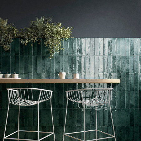 Azulejos verdes y brillantes forman un telón de fondo de pared debajo de plantas colgantes. Los taburetes de bar de metal blanco