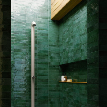 Ein vertikaler weißer Duschkopf steht vor glänzenden dunkelgrünen Fliesen in einer Dusche. Deckenpaneele aus Holz sorgen darüber