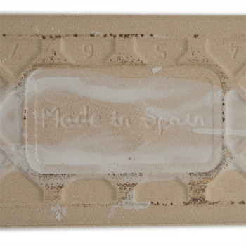 Eine rechteckige beige Fliese zeigt den geprägten Text „Made in Spain“, umgeben von einem strukturierten Muster mit den Zahlen 9