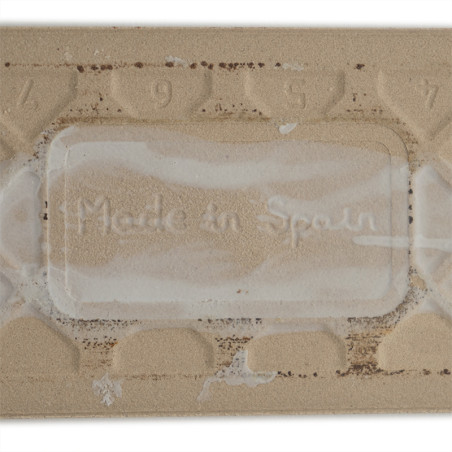Eine rechteckige beige Fliese zeigt den geprägten Text „Made in Spain“, umgeben von einem strukturierten Muster mit den Zahlen 9