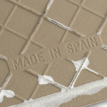 Un azulejo cerámico texturizado presenta el texto en relieve `MADE IN SPAIN` y está parcialmente cubierto con adhesivo blanco, c