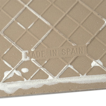 A beige ceramic tile featuring a grid pattern with white residue, displaying the text `MADE IN SPAIN` in an embossed style.