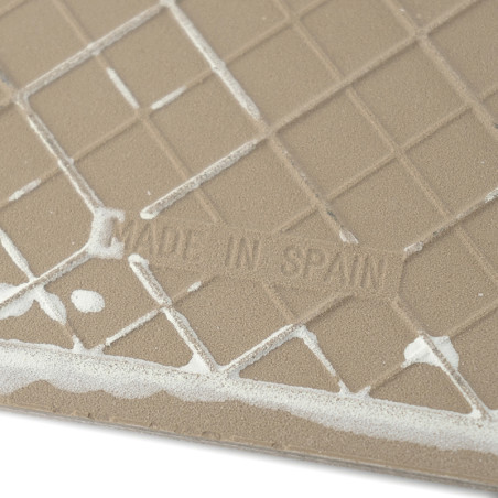 Eine beige Fliese weist ein geometrisches Gittermuster mit einigen weißen Rückständen auf. Der Text lautet „MADE IN SPAIN.“