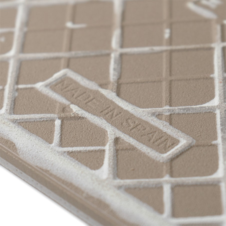 A beige tile features embossed, grid-like patterns. Raised text reads `MADE IN SPAIN.` The tile surface shows slight texture var A beige tile features embossed, grid-like patterns. Raised text reads `MADE IN SPAIN.` The tile surface shows slight texture var