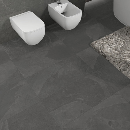 Des carreaux gris recouvrent le sol et les murs de la salle de bains, soutenant des toilettes et un bidet muraux. Un tapis à mot