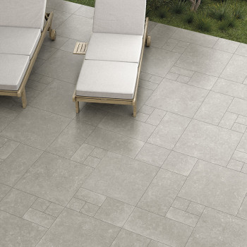 Los azulejos de piedra gris forman un patrón geométrico en un patio con dos sillones acolchados cerca, rodeados de césped y vege