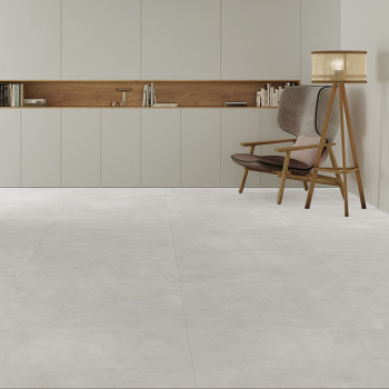 Una habitación minimalista cuenta con grandes azulejos de color gris claro. Una silla de madera con cojines y una lámpara de pie