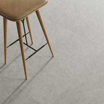 Auf einem hellgrauen Fliesenboden steht ein Holzhocker, dessen Beine Schatten werfen und so an einen minimalistischen Innenraum 