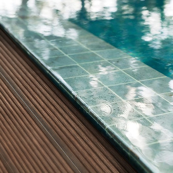 Une grille de carreaux de pierre turquoise borde une piscine réfléchissante à côté d’une terrasse en bois texturée.