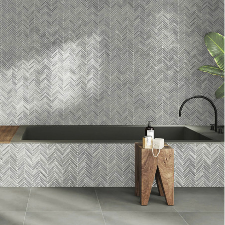 Graue Steinfliesen bilden eine Wand mit Fischgrätenmuster über einer Badewanne, ergänzt durch Holzhocker und eine Pflanze.