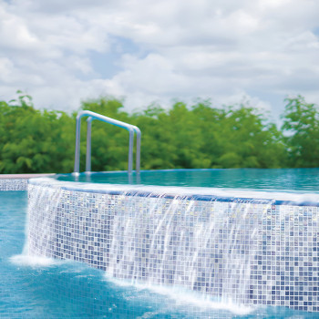 Cascatas de água sobre a borda de uma piscina infinita em mosaico de vidro, em meio a uma vegetação exuberante e um céu nublado.
