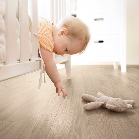 Un bebé se inclina sobre una cuna blanca, alcanzando un conejo de peluche sobre un piso de madera, en una habitación suavemente 