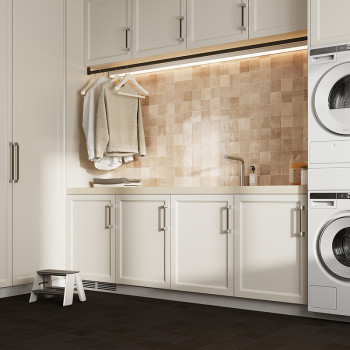 A laundry room features beige square tiles as a backsplash behind a sink, with white cabinets below. Clothes hang from a rod, an