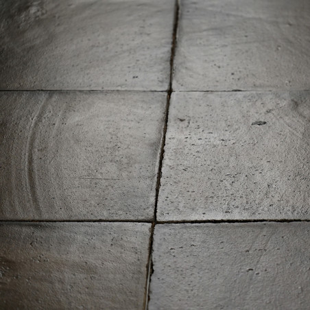 Des carreaux carrés en terre cuite sont adjacents, subtilement texturés avec des motifs circulaires, formant un sol patiné dans 