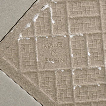 Eine beige Keramikfliese weist ein Gittermuster mit dem Text „MADE IN SPAIN“ in der Mitte auf, umgeben von hellen, diagonalen Fu