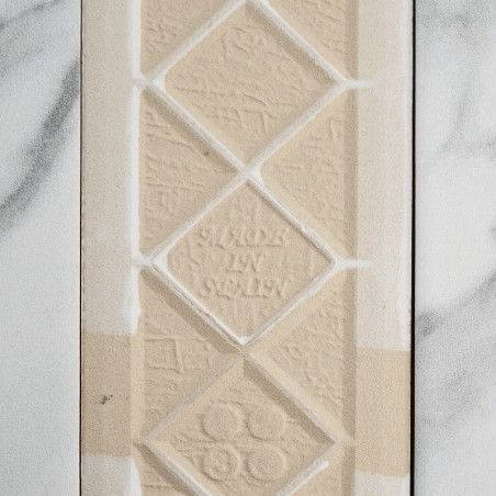 Un azulejo texturizado de color beige muestra formas de diamantes en relieve con el texto `MADE IN SWEDEN` en el centro. Se apoy Un azulejo texturizado de color beige muestra formas de diamantes en relieve con el texto `MADE IN SWEDEN` en el centro. Se apoy