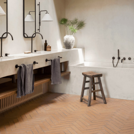 Terracottategels vormen een visgraatpatroon op de badkamervloer, met een houten krukje en een minimalistisch decor eromheen.