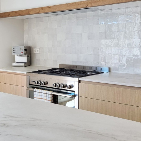 Glanzend witte terracottategels vormen een achterwand boven een moderne kachel in een strakke, minimalistische keuken. Glanzend witte terracottategels vormen een achterwand boven een moderne kachel in een strakke, minimalistische keuken.