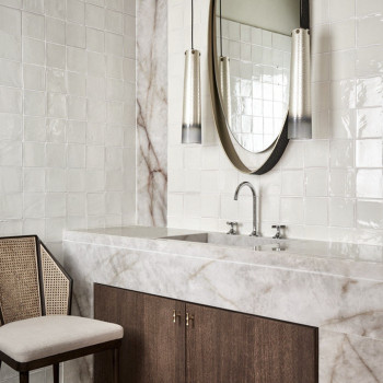 White terracotta tiles cover a bathroom wall, reflecting light. marble sink beneath, flanked by modern mirrors and lamps.