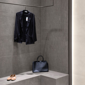 Un blazer noir est suspendu à un crochet dans un vestiaire minimaliste avec des carreaux gris foncé, à côté d'un sac à main noir