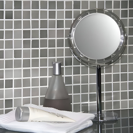 Circular mirror stands on a counter, reflecting a grid-patterned mosaic wall, beside a lotion dispenser and folded towels.