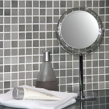 Le miroir se trouve à côté d’un flacon pompe et d’un tube de crème posés sur des serviettes, se reflétant sur un mur en mosaïque