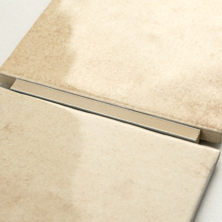 Two beige ceramic tiles rest side by side, separated by a narrow metal strip, on a smooth, neutral-colored background.