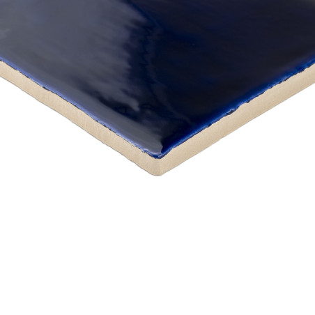 Un carreau de céramique brillant d’un bleu profond reflète la lumière, mettant en valeur sa surface vitrée lisse sur un bord bei