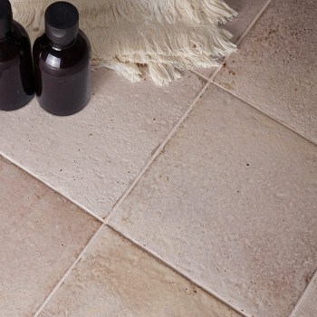 Duas garrafas escuras e tampadas estão sobre azulejos bege com superfícies texturizadas. Um pano esbranquiçado com franjas fica 