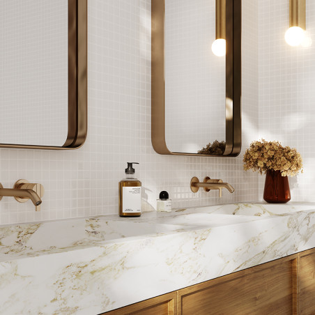 Des robinets muraux marron s'étendent sur un lavabo en marbre blanc veiné d'or. Deux miroirs se trouvent au-dessus, flanqués de 