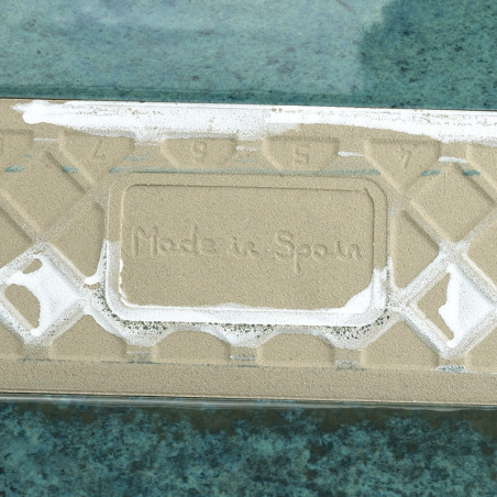 O ladrilho retangular, com a inscrição “Made in Spain”, fica plano, parcialmente coberto de areia, contra uma superfície texturi